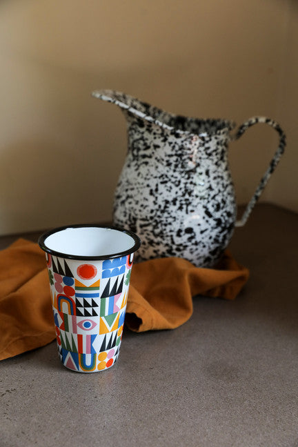 colorful metal tumbler set on a counter with a black spatterware pitcher and a dishtowel.