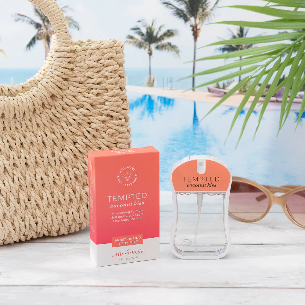 A Tempted Coconut Kiss moisturizing body mist bottle and its coral-pink packaging box displayed on a white wooden surface with a straw tote bag, sunglasses, and a tropical poolside background.