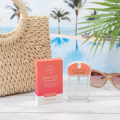 A Tempted Coconut Kiss moisturizing body mist bottle and its coral-pink packaging box displayed on a white wooden surface with a straw tote bag, sunglasses, and a tropical poolside background.