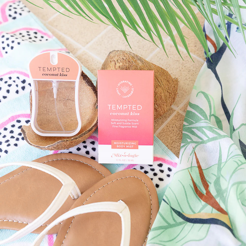 Tempted Coconut Kiss body mist packaging and bottle displayed on a beach-themed flat lay with tan sandals, coconut halves, palm leaves, and tropical-print fabric.