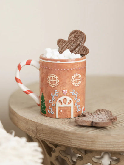 Gingerbread house mug set on a table and topped with marshmallows and a gingerbread cookie.