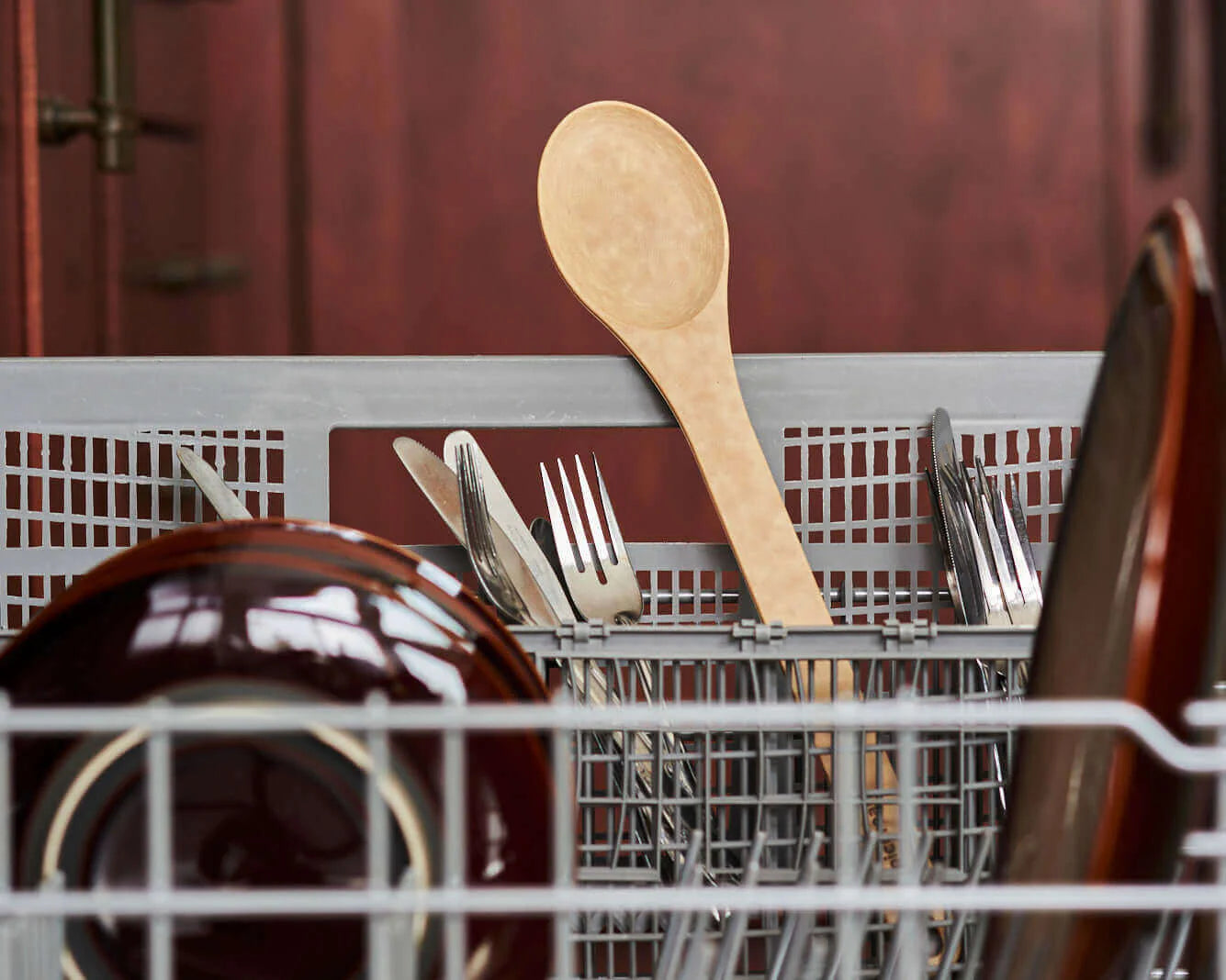 Dishwasher rack with dishes, including a wooden spoon and forks, against a blurred background.