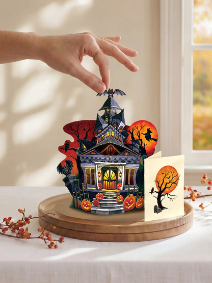 hand placing a small Halloween-themed pop up paper house on a round wooden base. a detailed haunted house, pumpkins, bats, a witch on a broomstick, and a separate card with a spooky tree and full moon, styled on a table near a window.
