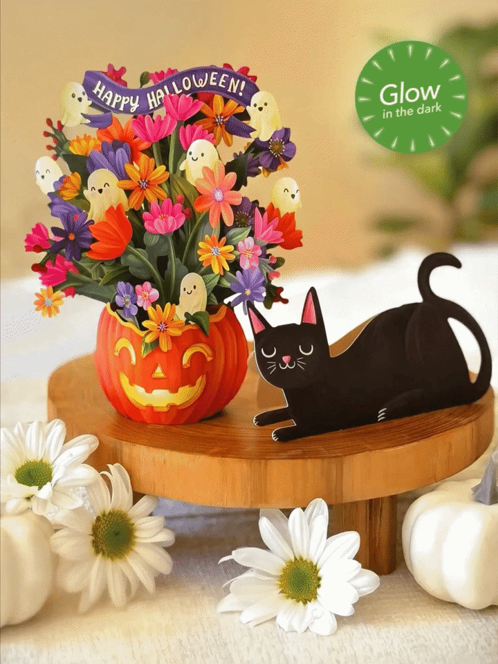 Halloween-themed decorative paper arrangement featuring a carved orange pumpkin vase filled with colorful artificial flowers and small ghost accents, alongside a black cat figurine on a wooden stand. White daisies and small pumpkins are placed around it, with a green 'Glow in the dark' badge in the top right.