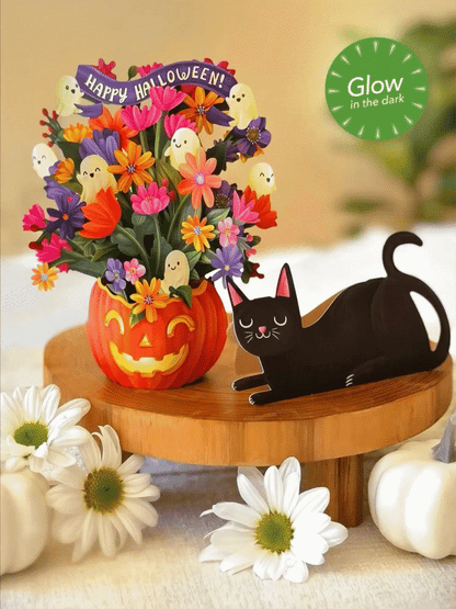 Halloween-themed decorative paper arrangement featuring a carved orange pumpkin vase filled with colorful artificial flowers and small ghost accents, alongside a black cat figurine on a wooden stand. White daisies and small pumpkins are placed around it, with a green 'Glow in the dark' badge in the top right.