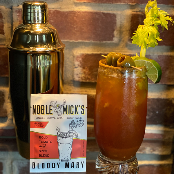 noble mick's single serve packet of bloody mary mix on a bar top with a cocktail shaker and a glass of bloody mary.