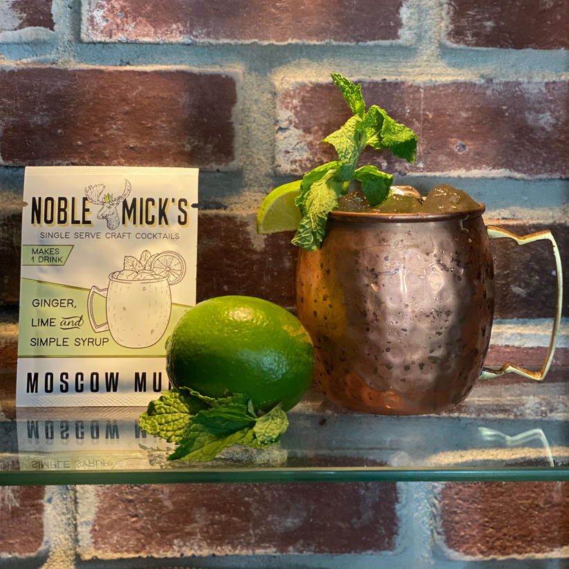 top view noble mick's single serve packet of Moscow Mule mix on a bar  with a  Moscow Mule, a lime, and mint sprigs.