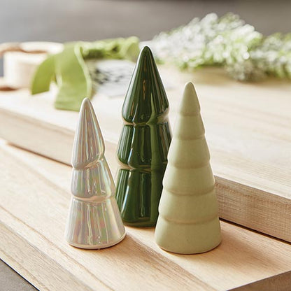 3 sizes and colors of ceramic trees set on a block of wood with greenery on the background.