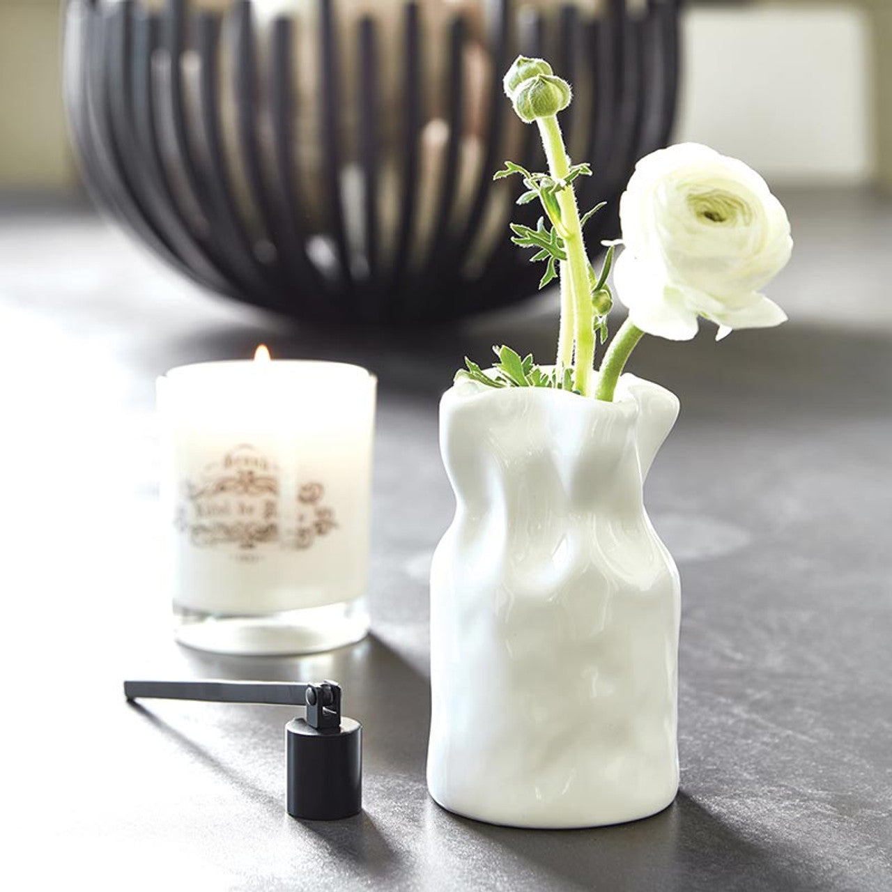 small cinched vase with a flower in it set on a desk with a candle and candle snuffer.