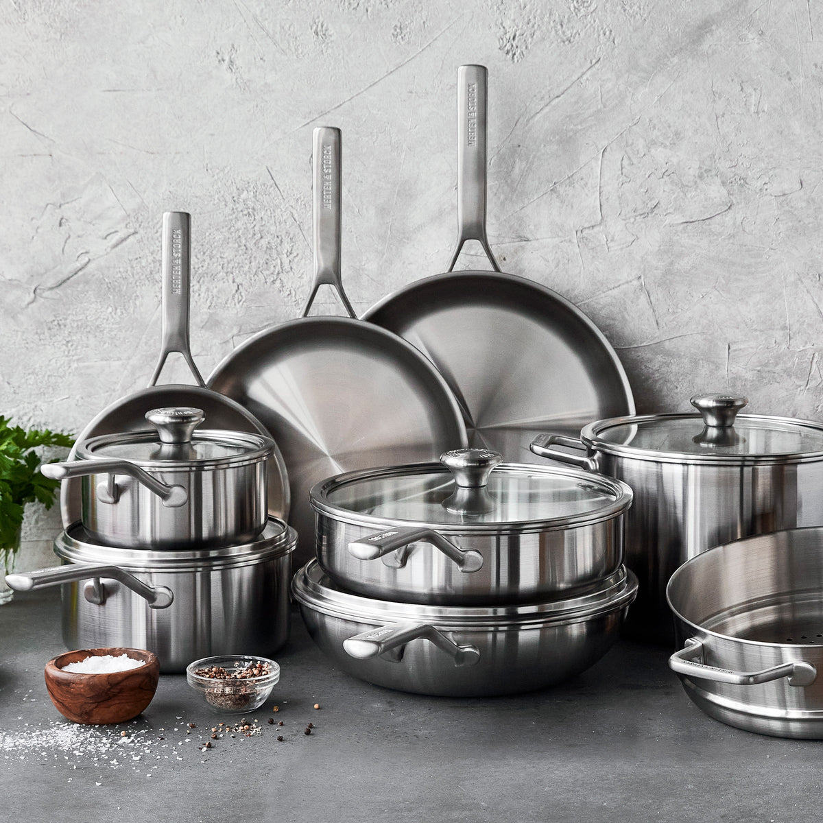 14 piece stainless steel cookware set arranged on a stone countertop with spices.