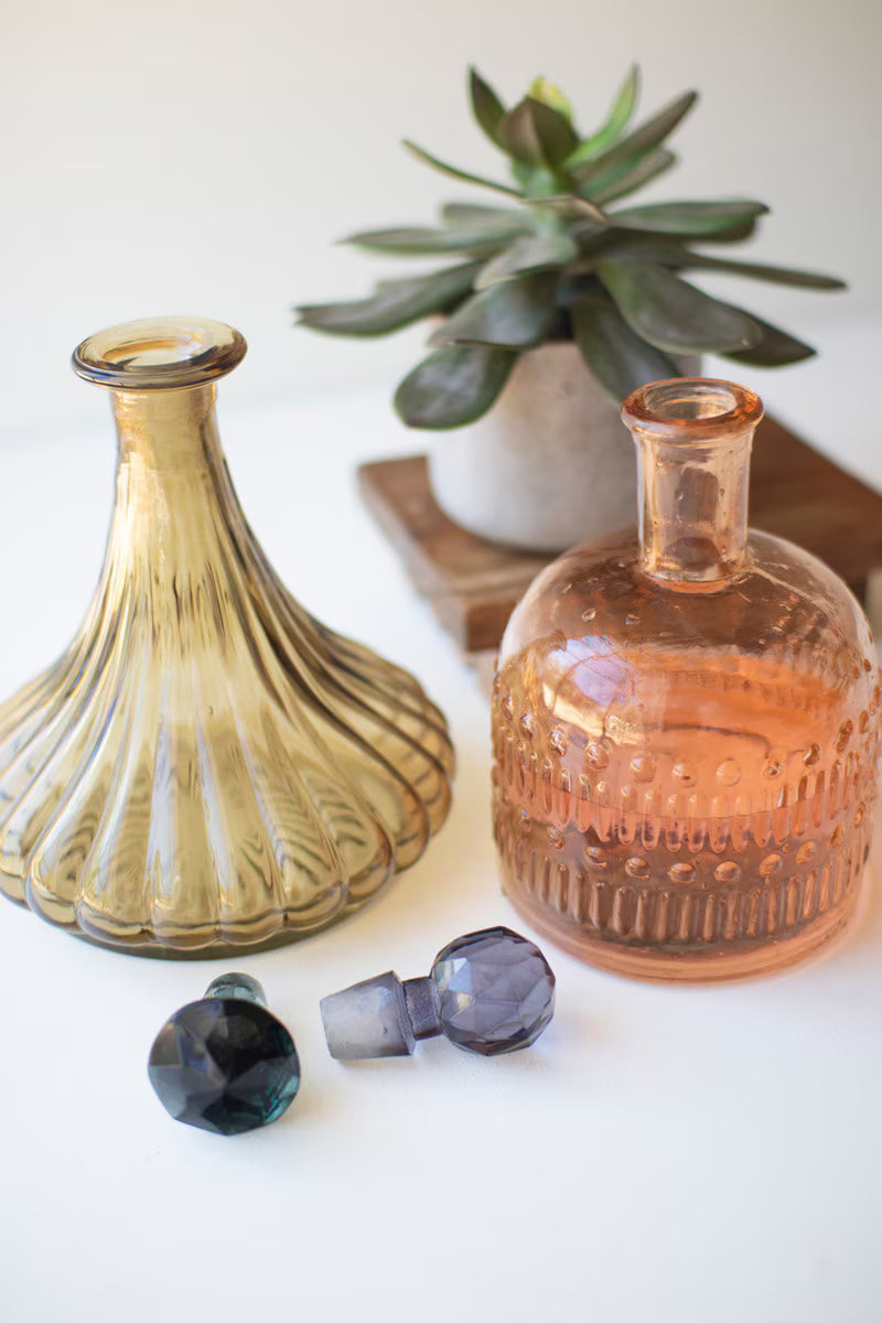 Two glass bottles with decorative caps on a white surface with a plant in the background.