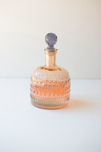 blush colored glass decanter with lavender stopper shown on a white background