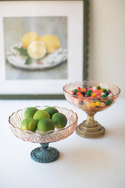 Two glass compotes, one smaller with green limes and one larger with colorful candies, against a white background with a framed picture on the wall.