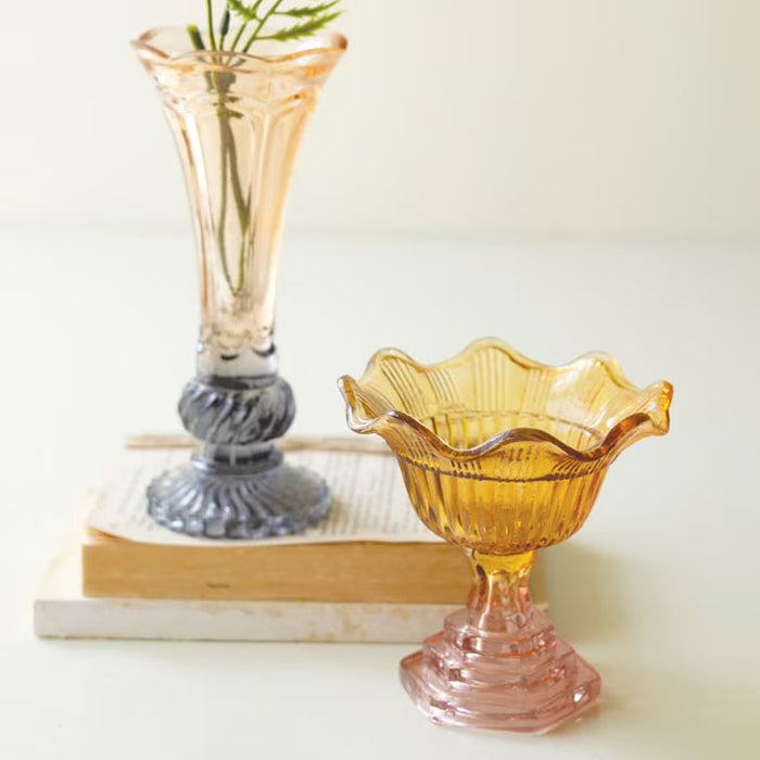 Two glass vases, one pink and one amber, on a white surface with books in the background