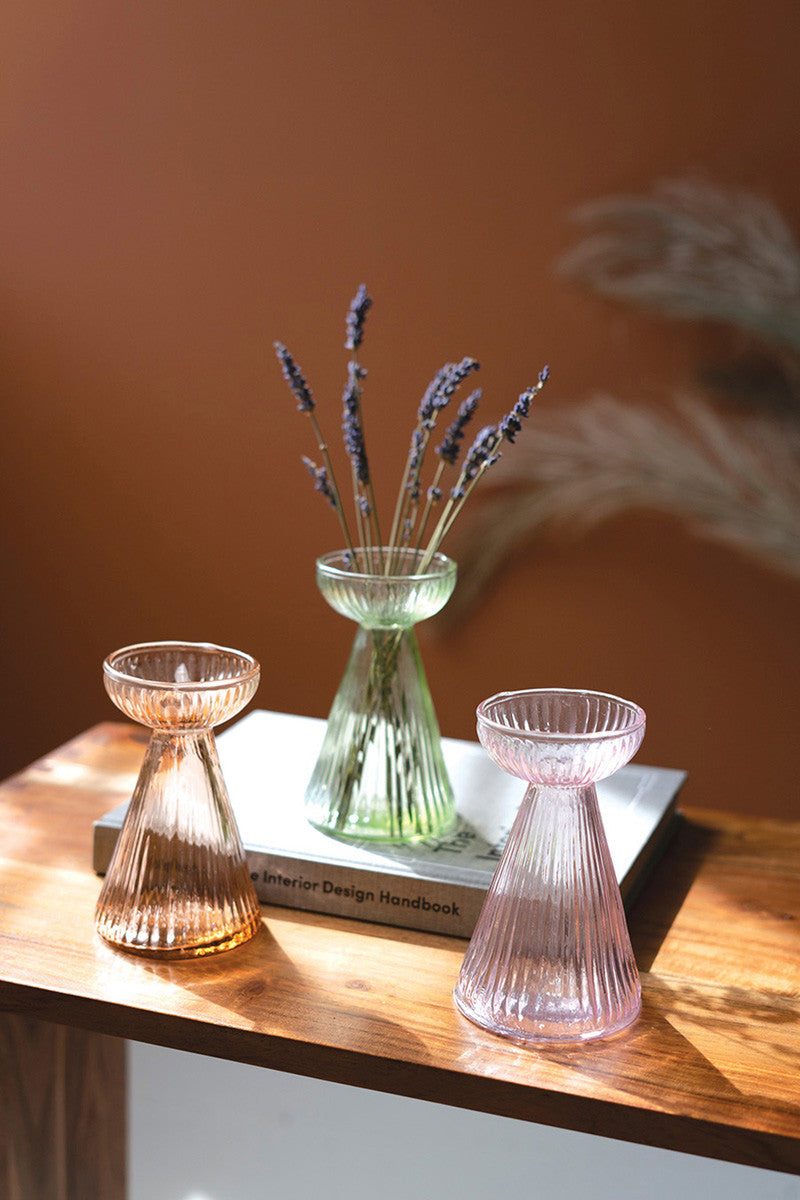 3 colors og glass vases set on a wooden table, one of the vases in set on a book and has lavender in it.