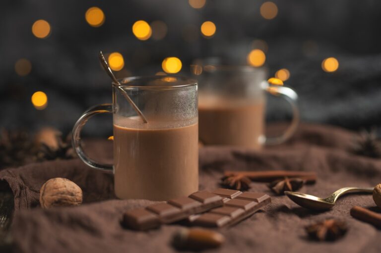 2 glass mugs filled with hot chocolate set on a table with a towel, bits of chocolate, and spices.