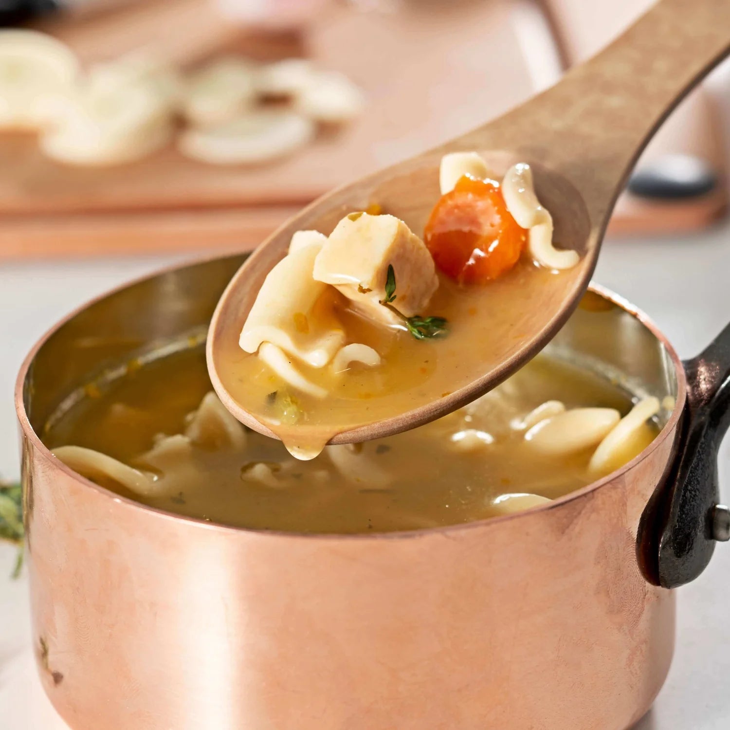 Copper pot with chicken noodle soup and a wooden spoon lifting a scoop of soup.