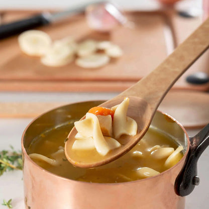 Copper pot of soup with a wooden spoon lifting noodles and carrots.