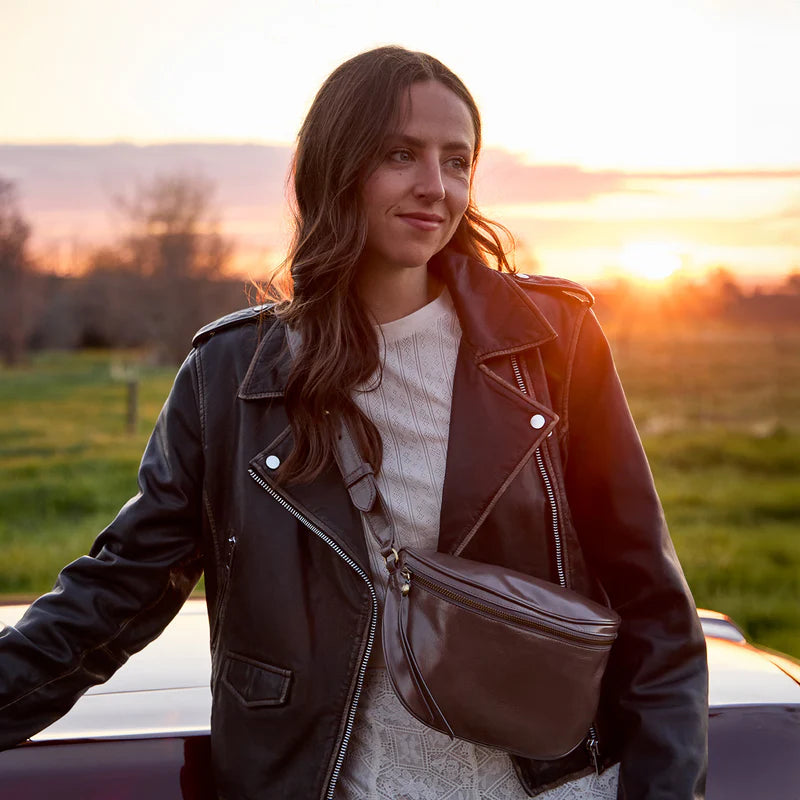 Person wearing black leather jacket standing outside with Hearthstone Juno belt bag over their shoulder and the sunset in the background
