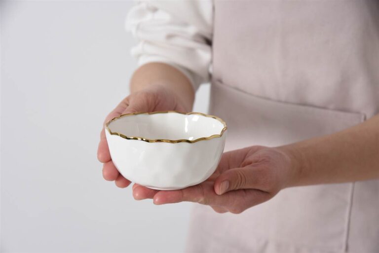 Person holding a white ceramic bowl with gold rim against a plain background