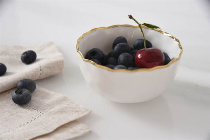 White bowl with gold rim filled with blueberries and a cherry on a white surface with a beige cloth.