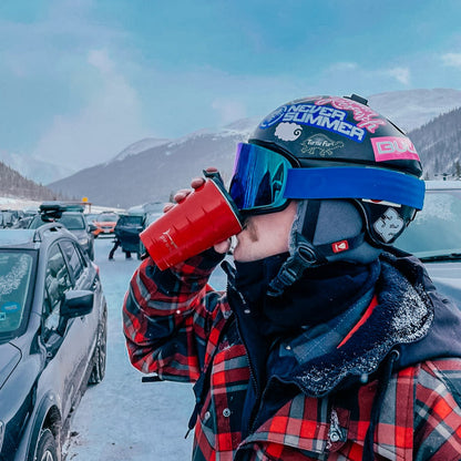 person wearing ski gear and sipping from a red tumbler in a parking lot with snow covered mountains in the background.