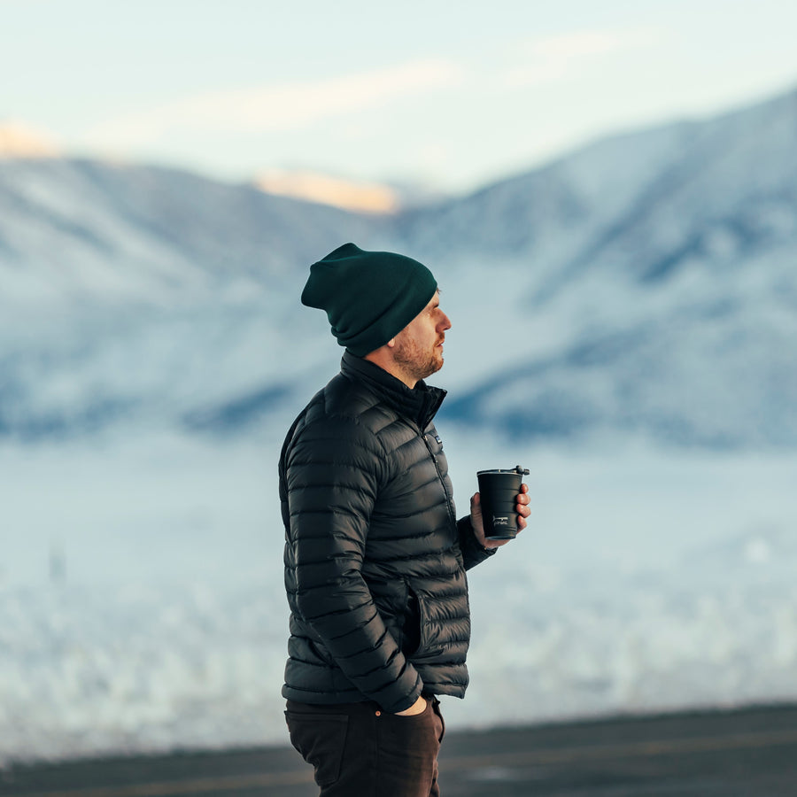 person wearing a jacket and hat holding a black tumbler with snow covered mountains in the background.