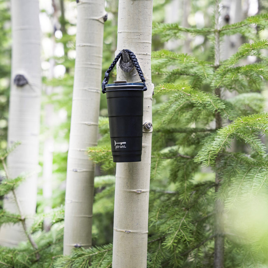 black tumbler with silver pirani logo near the bottom of cup with a lid on it hanging on a tree branch by a braided handle.