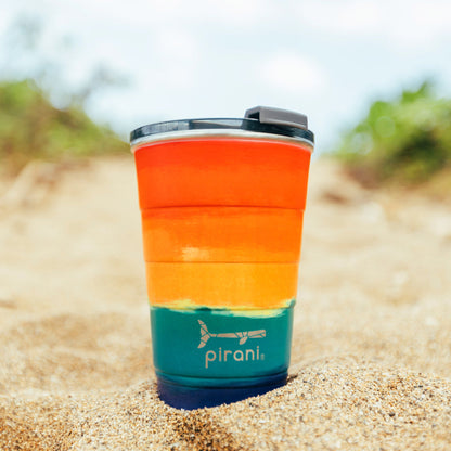 sunset tumbler with silver pirani logo near the bottom of cup and a lid on it set in the sand with beach in the background.