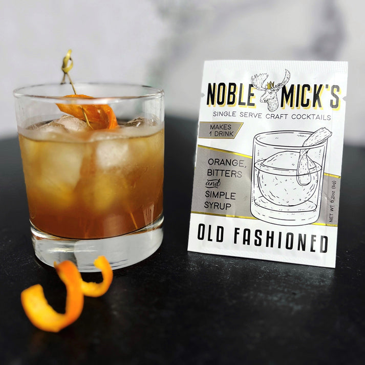 top view noble mick's single serve packet of Old Fashioned mix on a bar  with an Old Fashioned and an orange peel.