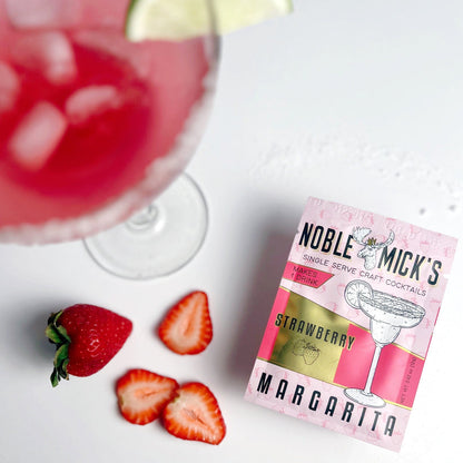 top view noble mick's single serve packet of strawberry margarita mix on a bar with a margarita and strawberries.