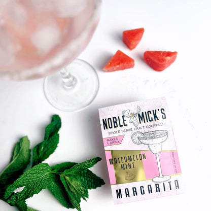 top view noble mick's single serve packet of Watermelon Mint margarita mix on a bar with a margarita, watermelon pieces and mint.