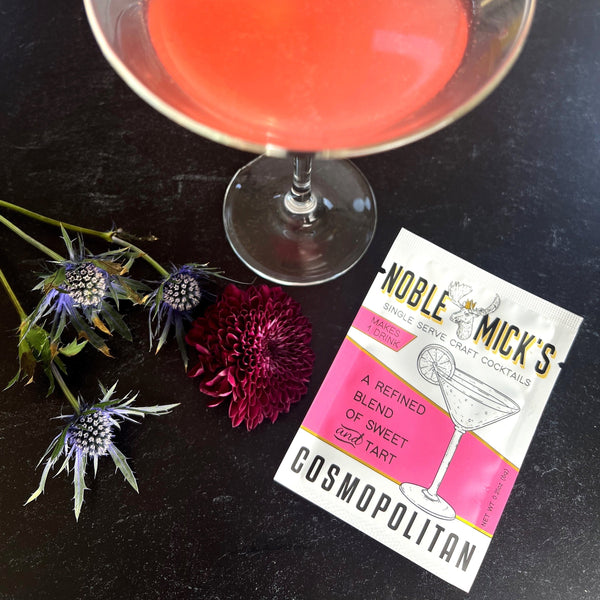 top view noble mick's single serve packet of cosmopolitan mix on a bar top with a glass of cosmopolitan and flowers.