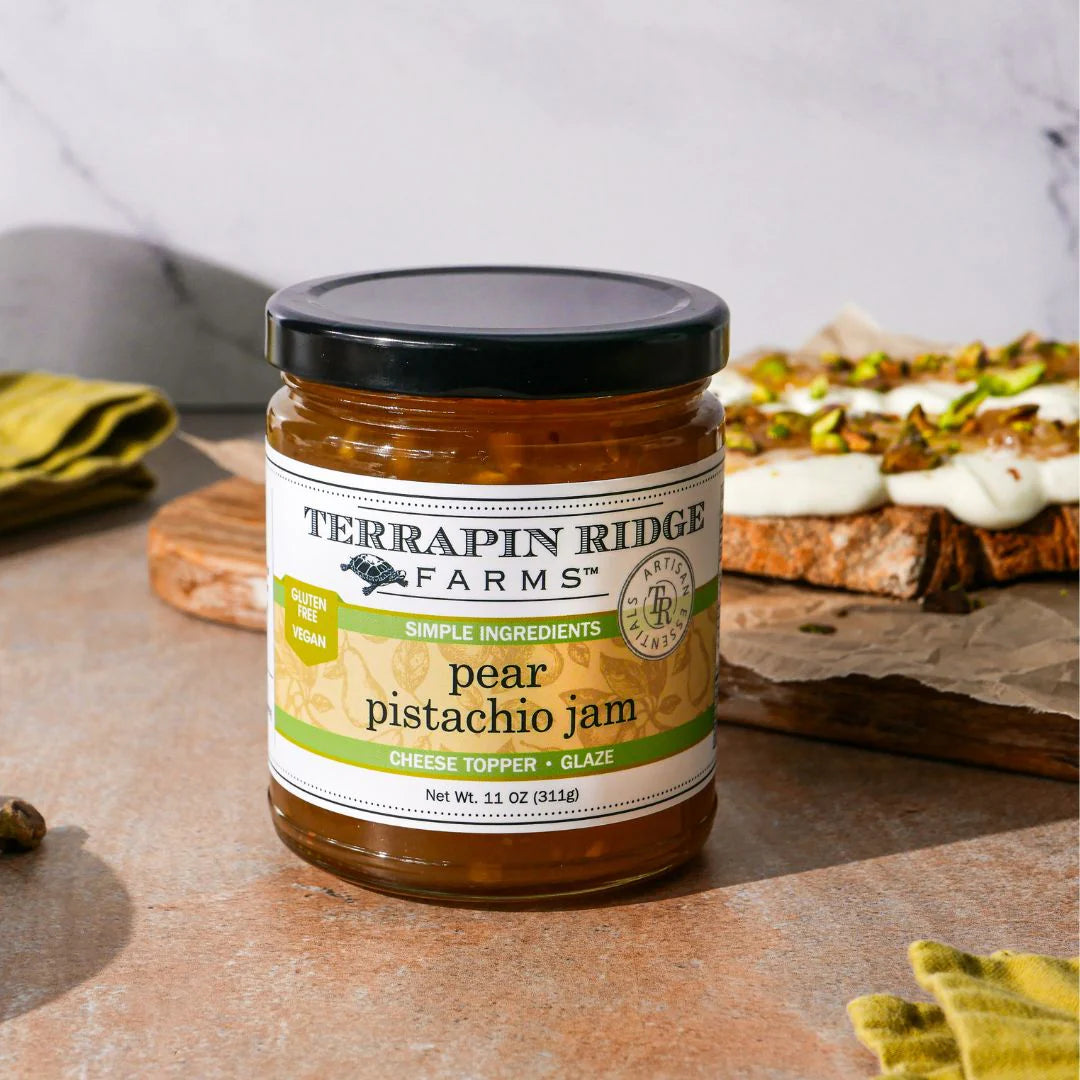 Jar of Terrapin Ridge Farms pear pistachio jam on a wooden surface with bread and cheese in the background.