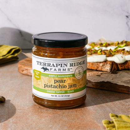 Jar of Terrapin Ridge Farms pear pistachio jam on a wooden surface with bread and cheese in the background.