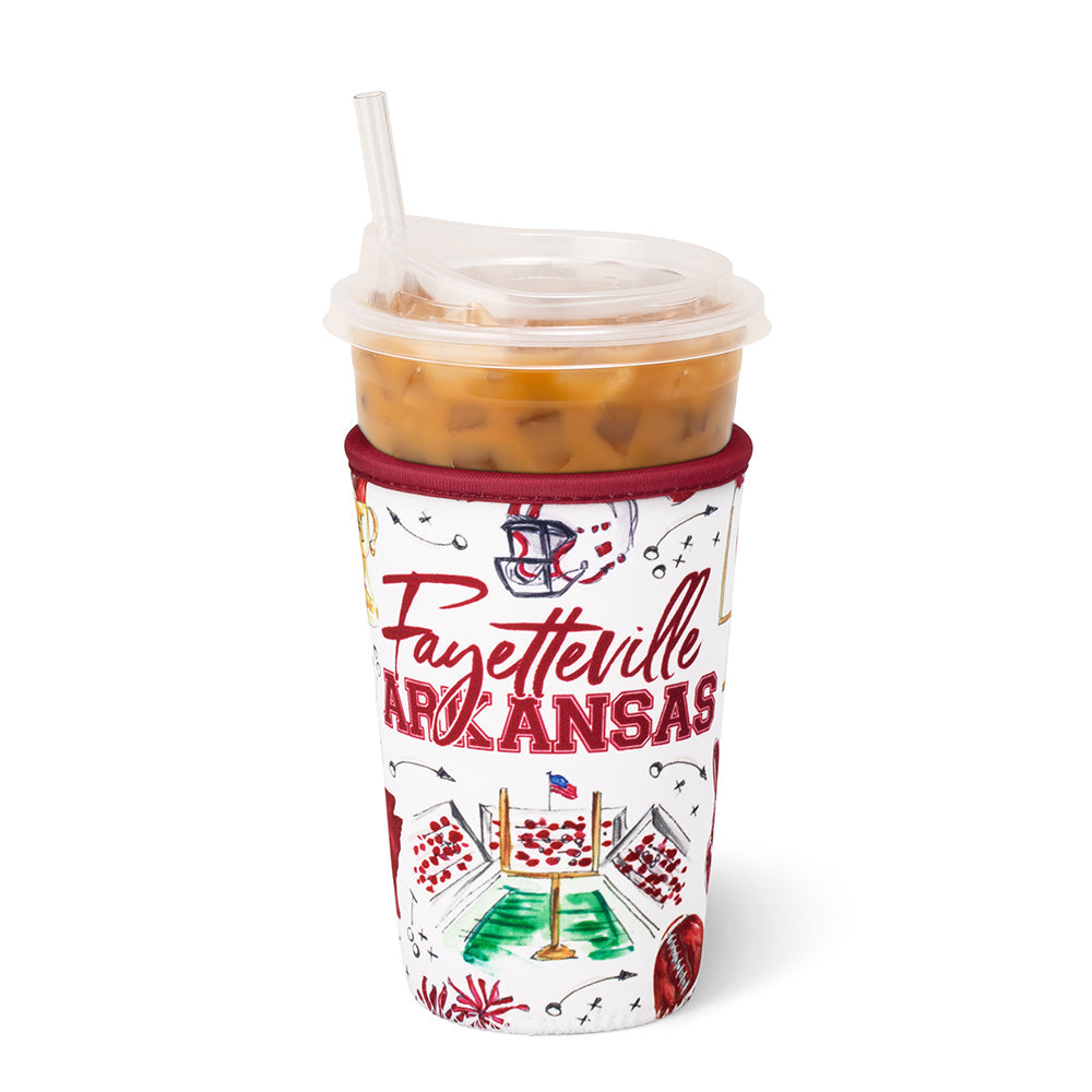 An iced coffee in a clear plastic cup with a clear lid and straw, in a white fabric sleeve printed with 'Fayetteville Arkansas' in red script and sports-themed graphics, on a white background.