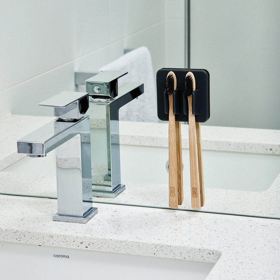 black silicone toothbrush holder with toothbrushes in it on a mirror by a bathroom faucet.