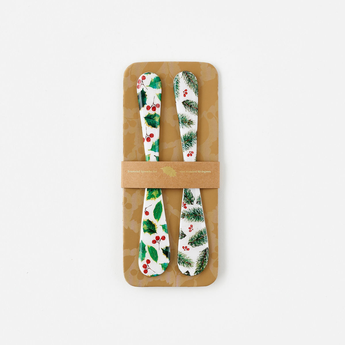 Two decorative spreaders with a holly and pine designs in a cardboard holder on a white background