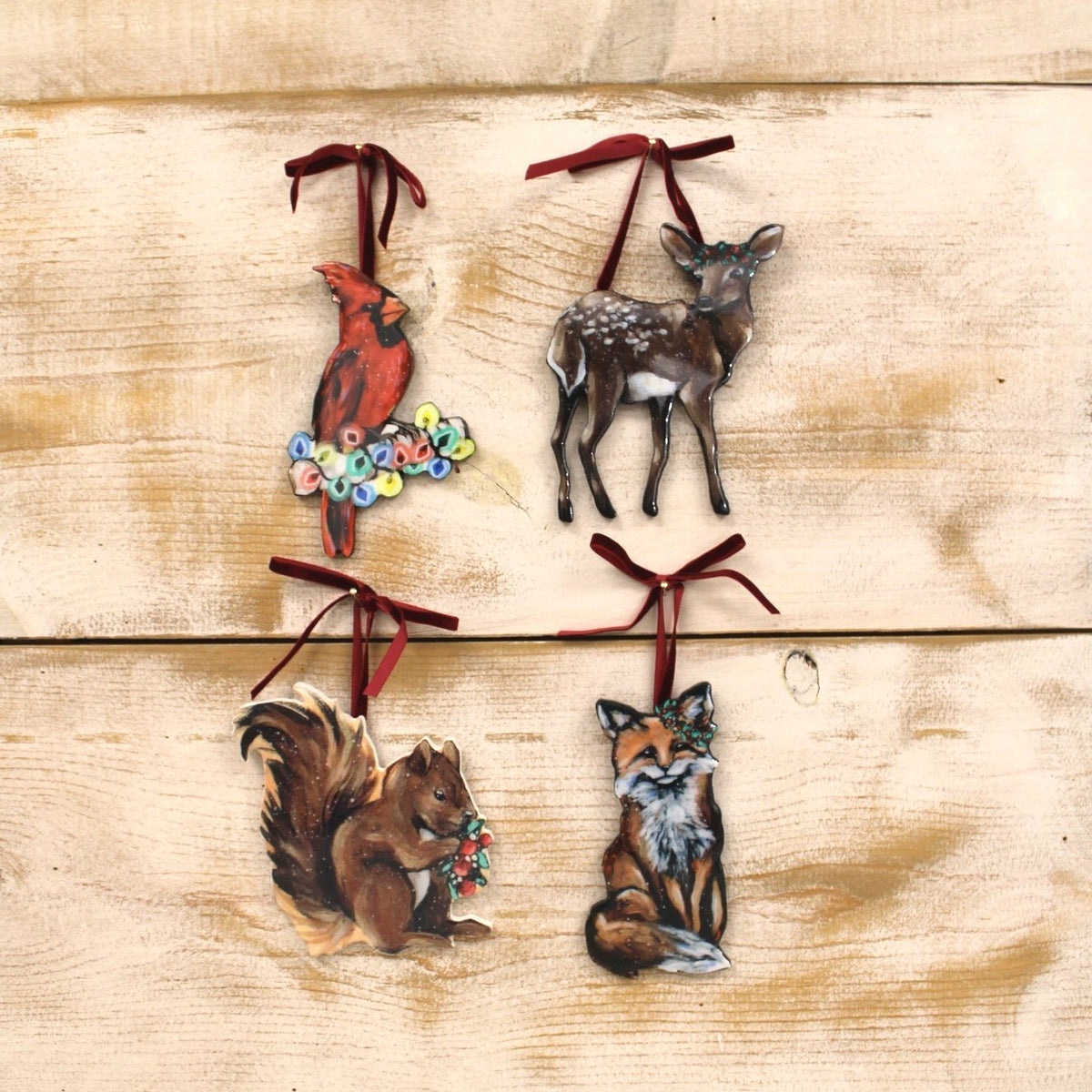 Four Christmas tree ornaments shaped like a cardinal, deer, squirrel, and fox, each with a dark red ribbon hanger, displayed on a light wood background.