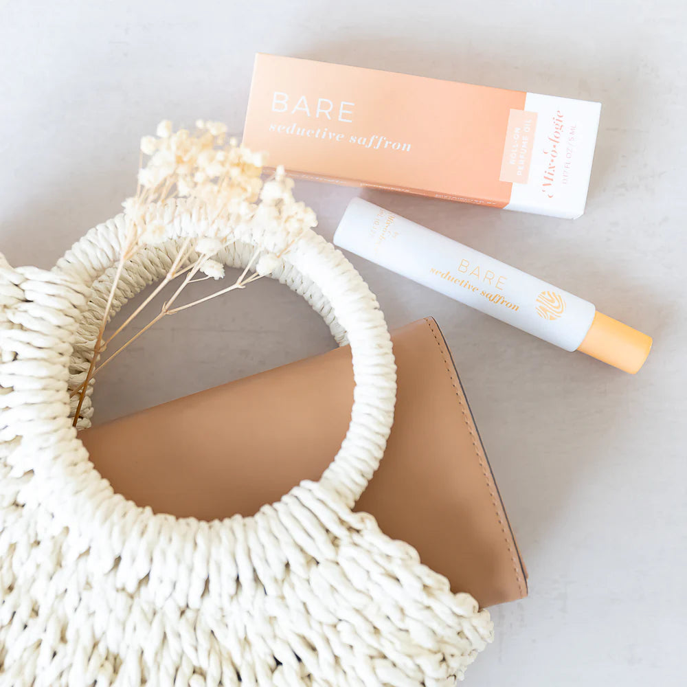perfume and its packaging arranged on a grey surface with a braided bag and dry botanicals