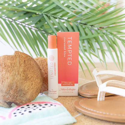Tempted Coconut perfume and box displayed on a beach-themed surface with a coconut, white sandals, and green palm leaves in the background.