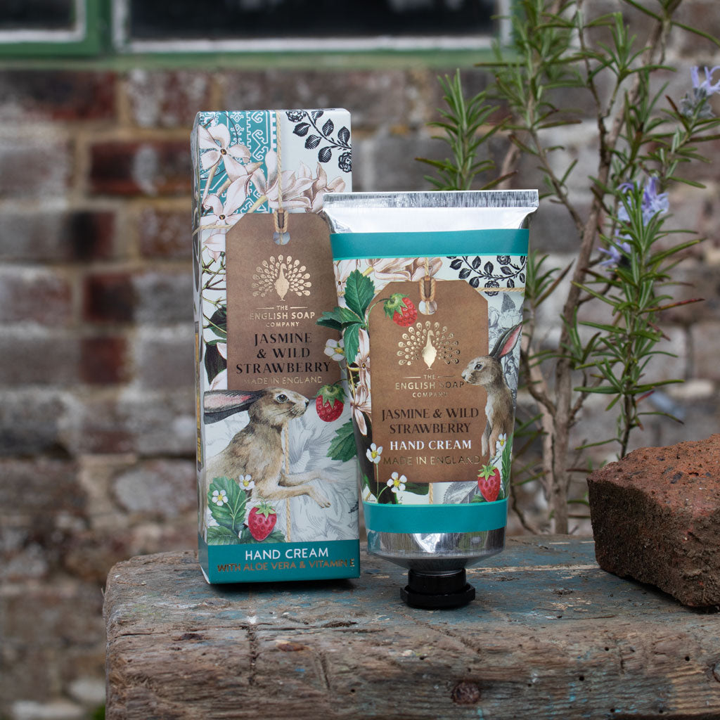Hand cream and its box packaging with floral and berry designs on a wooden surface against a brick wall.