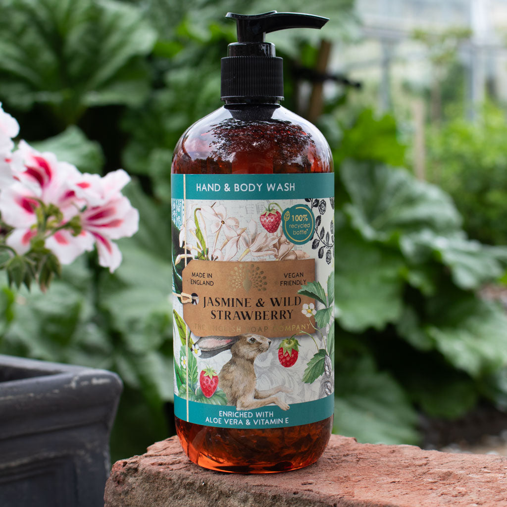 Hand and body wash bottle with a label featuring jasmine and wild strawberry on a wooden block with plants in the background.