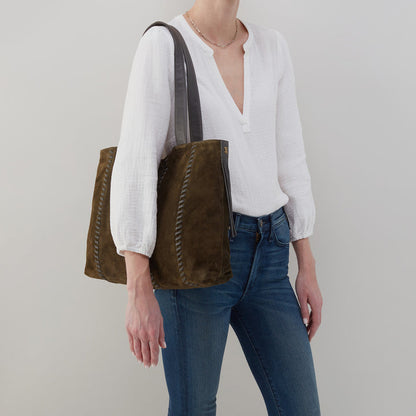 person wearing jeans and a white blouse carrying a dark green suede tote bag on their shoulder.