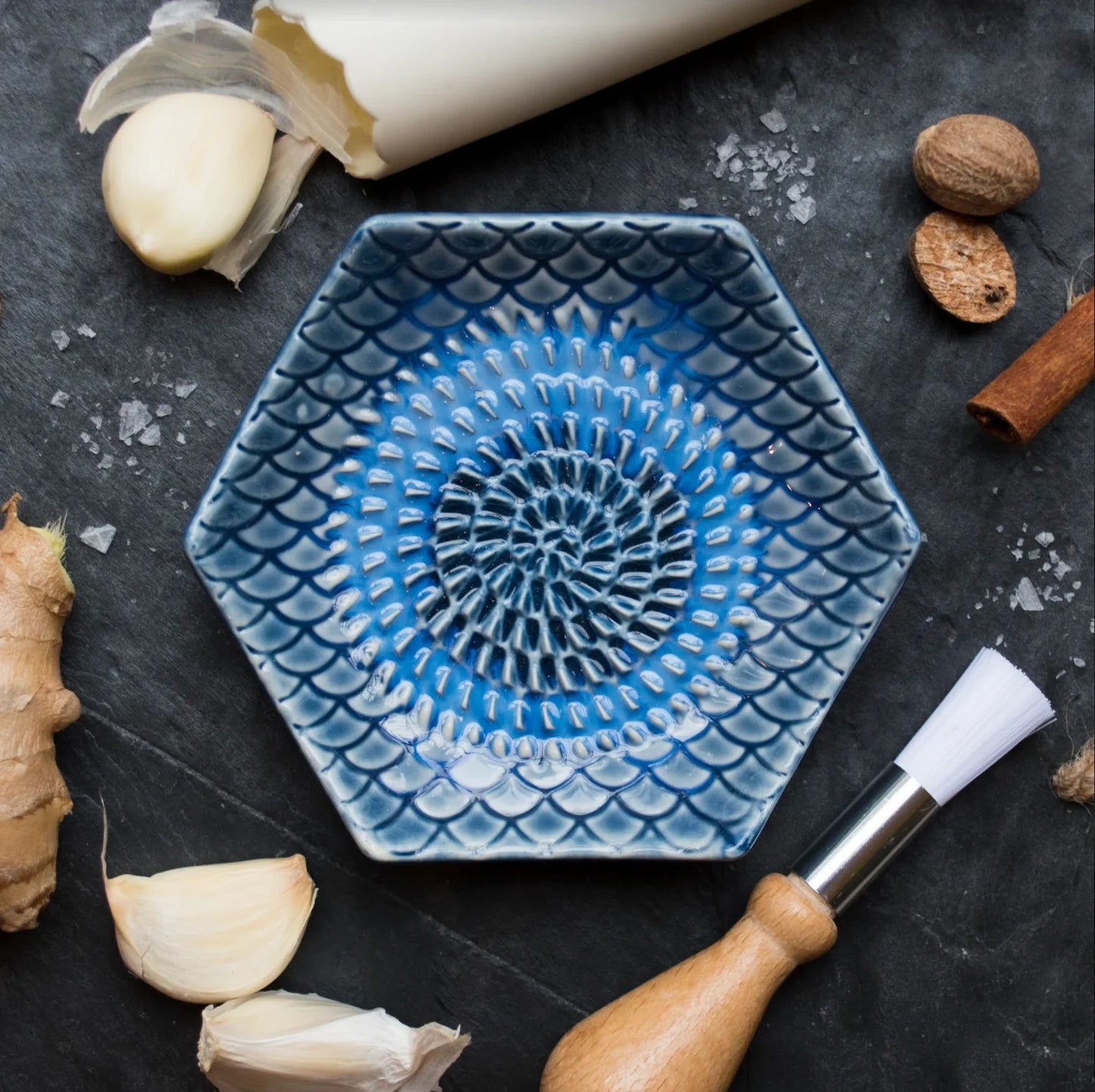top view of a black countertop arranged with the blue tie dye grate plate, silicone garlic peeler, brush, and a variety of spices and roots.
