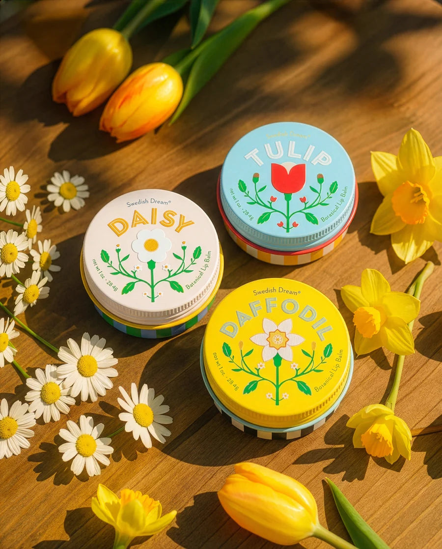 Three assorted tins of floral lip balm, arranged on a wooden surface surrounded by flowers.