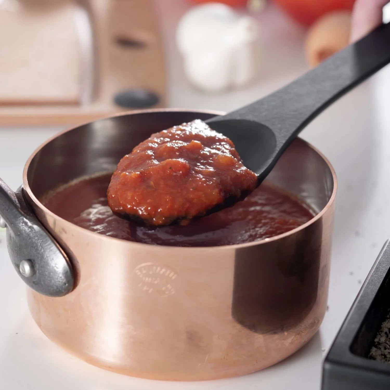 Copper pot with a spoonful of red sauce being lift by a black spoon on a kitchen counter