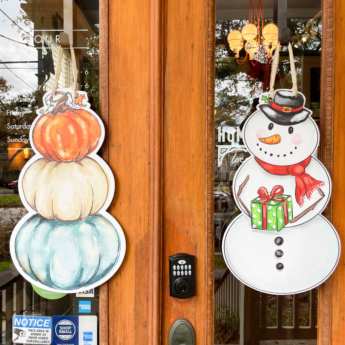 Decorative door hangers, one of pumpkins and the other a snowman, both hung on a wooden and glass door.