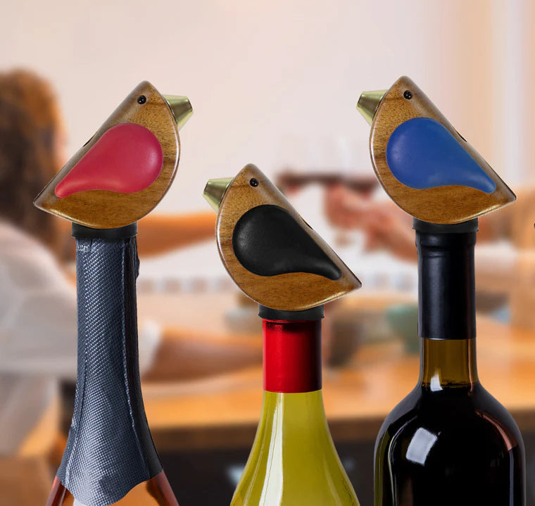 3 bird-shaped bottle stoppers on wine bottles with a blurred background of people and a table.