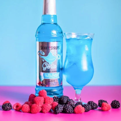 bottle of sour genie skinny syrup set next to a stemmed glass of a blue beverage and surrounded with raspberries.